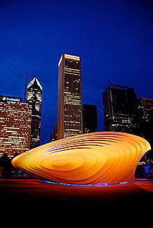 an amber structure with spectators milling about and a backdrop of tall buildings