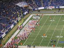 Several American football players in purple and orange uniforms standing in the end zone of a stadium. Spectators are visible on the left and top sides.