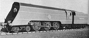 A posed side-and-front view of a large 4-6-2 steam locomotive with a tender. The locomotive boiler is hidden by a casing of flat metal side sheets.