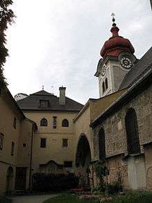 Part of a partially planted courtyard. As viewed, on the western and northern sides is a four storey building with a smooth yellow render and dark doors and windows; on the eastern side is an archway surrounded with the same render, with the remainder of the building face in stone with dark windows. Above the arch is a square, white clocktower with a red, octagonal, bulbous spire.