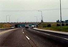 Driving down a six-lane highway during the day. In front is a concrete bridge. The highway curves to the right as it passes beneath the bridge.