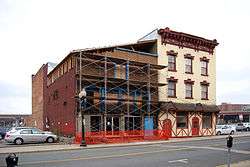 Two attached buildings, with a large parking lot around and behind them and an elevated highway in the background. The one on the right is brick, three stories high, painted yellow with an ornate decoration at the flat roof. On the left is another brick building with a sloping roof and scaffolding in the front.