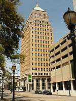 Ground-level view of a 20-story building with a tan brick facade; the roof of the structure is a silver pyramid that tapers into a flagpole.