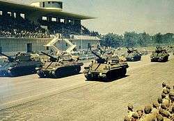 Colour picture of Sherman tanks passing a saluting diaz in an autodrome.