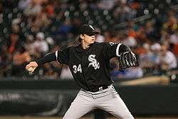 A young man in a black baseball jersey and cap with white trim and white baseball pants throws a baseball with his left hand.