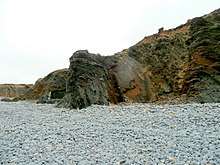 a cliff of folded grey and brown rocks, looming over a beach of grey cobbles