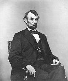 Middle-aged man in a beard posed sitting in a suit, vest and bowtie