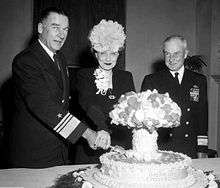 A man in naval uniform and a woman wearing a hat cut into a cake labeled Operation Crossroads, and shaped like a mushroom cloud, while another naval officer looks on.