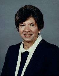 A color photograph portrait of a woman wearing black clothing with a white collar and white border, single pearl earrings, her thick dark hair set in a conservative bouffant style, smiling and looking at the camera