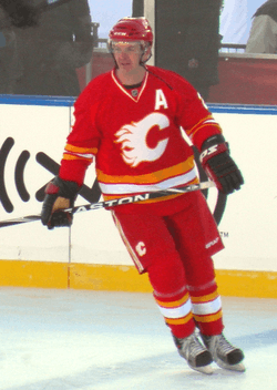 An ice hockey player looks to his right as he glides across the ice.  He is in a red uniform with white and yellow trim, with a stylized "C" logo on his chest.