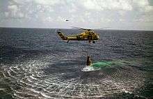 A green helicopter hovers low over the water, with the Mercury capsule suspended below. The helicopter has "Marines" written on it, and the number "44"