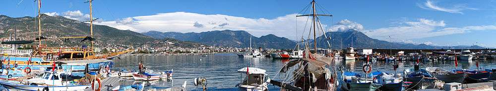 A panoramic view of the east side of the city from the old harbour near Kızılkule. Numerous white, blue, and orange boats float tied to a dock in a greenish sea. Mountains rise on the other side of a bay.