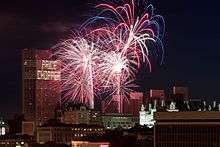 Blue and red fireworks explode over a complex of buildings after dusk.