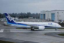 B787 in launch customer All Nippon Airways' blue and white livery. In the background are two assembly halls, with huge doors facing left. Vehicles are parked in front of the halls.