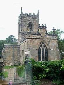 A stone church seen from the east.  It has a three-light east window, over which is a sundial, and beyond it is a battlemented tower