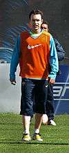 A man wearing long, dark blue shorts and a light blue sweatshirt under an orange bib.