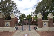 A brick wall with a gap in the middle. Through the gap one can see a statue at the bottom of a palm-lined walkway.