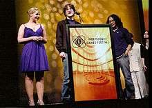 Two men stand behind a podium marked Independent Games Festival, while a woman in a blue dress stands to the left.