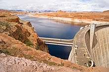 Side view of a large concrete dam impounding a lake surrounded by red-rock hills