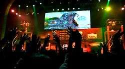 Performers on stage in front of large screen, seen from crowd with raised hands