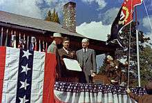 Men in suits and uniforms stand on a dais decorated with bunting and salute.