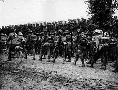 Ranks of marching men wearing Brodie Helmets and 1908 pattern webbing, rifles slung being watched in the background by Australian soldiers wearing Australian slouch hats