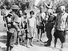 Black and white photo of three men wearing military uniforms and carrying guns with three elderly men and a teenager wearing civilian clothes