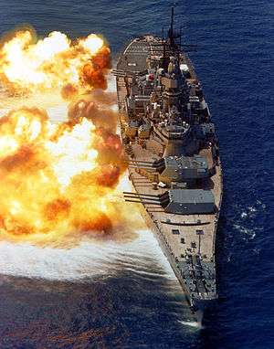 USS Iowa in her post 1980s rebuild. The battleship is pointed toward the viewer, with her 9 gun barrels pointed to the left for a gunnery exercise. Fire can be seen erupting from the gun barrels, and a concussive effect is visible on the water.