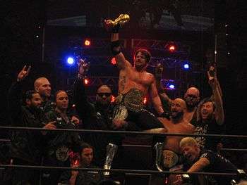 Ten members of the Bullet Club group celebrate in a professional wrestling ring. Several are using a hand gesture, extending the index and little finger, with the tips of the renaming fingers touching.