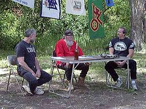 Three middle-aged white men sit at a camping table outdoors.