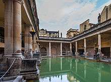 Photograph of the Baths showing a rectangular area of greenish water surrounded by yellow stone buildings with pillars. In the background is the tower of the abbey.