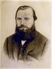 A man in his late 20s or early 30s with dark hair and a bushy beard, wearing a dark coat, dress shirt and tie