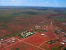  an aerial view of a small settlement, showing about seventy rooftops, red dirt roads and a dirt oval, with dry scrubland receding into the distance