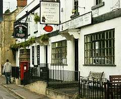 White fronted buildings with windows with small panes of glass. Shop signs for fish and chips and a pub. Postbox on the pavement in front of the buildings separated by black railings.