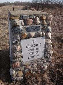 Stone cairn erected in 1975 marking the Battleford Industrial School Cemetery. A plaque at the top of the cairn reads: RESTORATION THROUGH OPPORTUNITIES FOR YOUTH, 4S1179-1974. PLAQUE PROVIDED BY DEPARTMENT OF TOURISM AND RENEWABLE RESOURCES. A