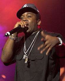 An African-American man, wearing a black shirt, black baseball cap and two neckchains, raps into a microphone.