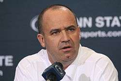 A man in a white shirt (Penn State coach Bill O'Brien) sits in front of a blue background with the words "Penn State" partly visible. He is addressing the media in front of a microphone emblazoned with the Penn State logo.