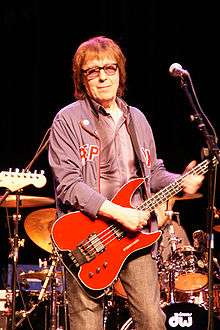 Musician Bill Wyman stands onstage beside a microphone, holding a bass guitar.