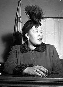 African-American woman, in her late thirties, sitting in a courtroom, looking worryingly to the right. She is wearing a feather hat, a dark coat, and a large ring on her right hand resting upon the witness stand.