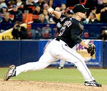 A man wearing a black baseball jersey and cap and white baseball pants throwing a baseball with his left hand