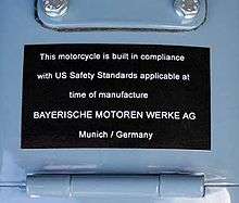 Black placard mounted on a light blue fuel tank, which reads "This motorcycle is built in compliance with US safety standards applicable at the time of manufacture BAYERISCHEN MOTOREN WERKE AG Munich / Germany"