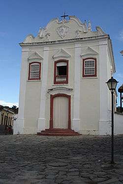 Facade of a two-storied stone church, largely painted white/beige with red/brownish window frames.