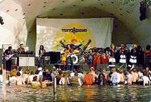 A crowd of people standing in water and listening to a band perform on stage