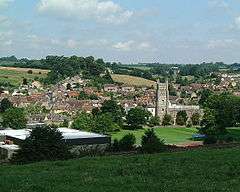 multiple buildings including a square church tower amongst fields and trees.