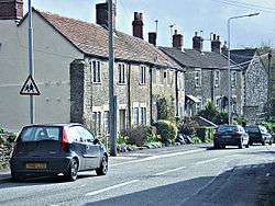 Street scene showing grey stone houses on the left of a road with a few cars.