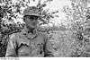 A black-and-white photograph of a man wearing a military uniform, field cap and a neck order in shape of an Iron Cross standing in front of a tree.