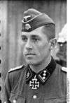 A man wearing a military uniform, side cap and neck order in the shape of a cross. His cap has an emblem in shape of a human skull and crossed bones.