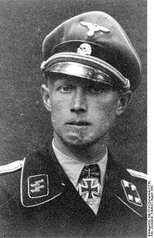 A black-and-white photograph of a man wearing a military uniform, peaked cap and a neck order in shape of an Iron Cross. His cap has an emblem in shape of a human skull and crossed bones. A large scar on his chin is visible.