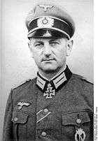 The head and shoulders of a man. He wears a peaked cap and a military uniform and an Iron Cross displayed at the front of his uniform collar. His facial expression is determined; his eyes are looking into the camera.