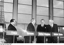 Four men stand behind podiums with their country names of France, Germany, United Kingdom, and United States, in front of a backdrop of the Eiffel Tower.